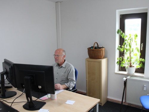 Aktuelles 82 Älterer Mann arbeitet an einem Computer in einem modernen Büro mit Pflanzen und natürlichem Licht.