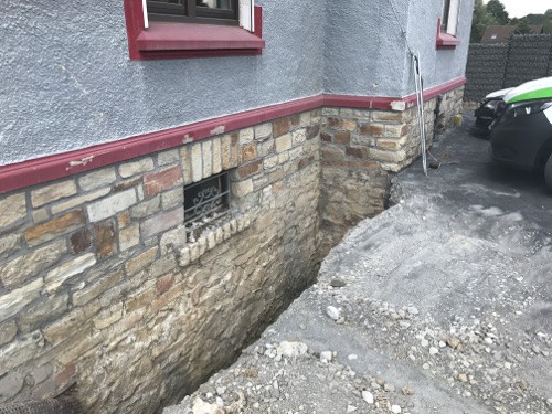 WIR ZIEHEN UM … 4 Graben entlang der Hauswand mit sichtbarem Mauerwerk und einem Fenster, das teilweise freigelegt ist. Der Boden ist uneben und zeigt lose Steine und Erdmaterial.