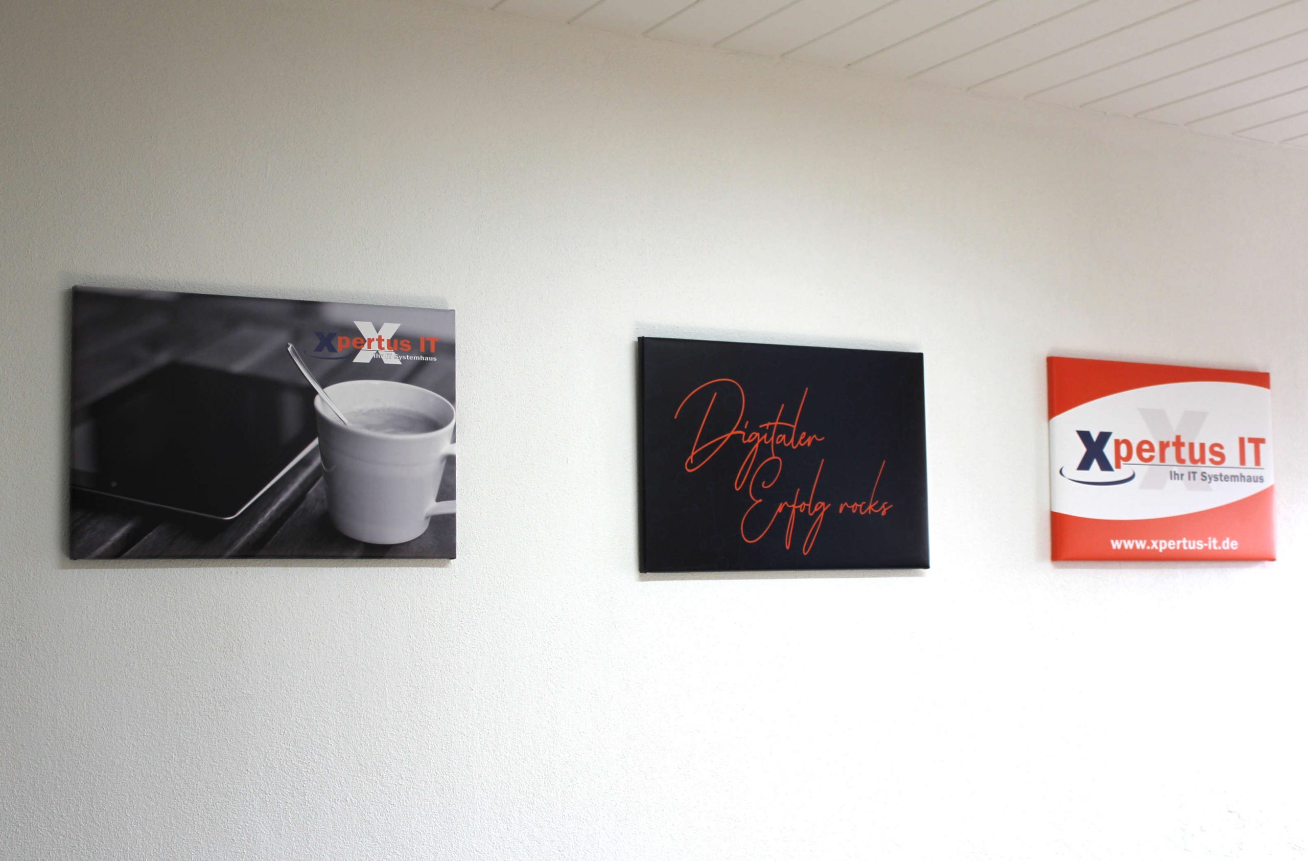 History of Xpertus 28 Drei Wandbilder im Büro von Xpertus IT: Links ein Bild mit einem Tablet und einer Tasse Kaffee, in der Mitte ein schwarzes Schild mit der Aufschrift „Digitaler Erfolg rocks“ und rechts das Logo von Xpertus IT mit Website-Adresse.