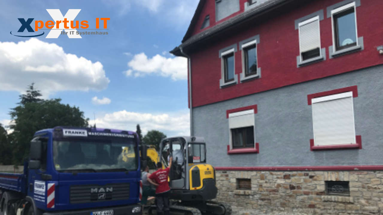 History of Xpertus 26 Bauarbeiter stehen neben einem blauen MAN-Lkw und einem gelben Minibagger vor einem renovierten Haus mit roter Fassade. Der Himmel ist blau mit einigen Wolken.