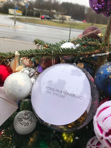 Weihnachtsbaum mit bunten Kugeln, darunter eine transparente Kugel mit dem Logo des Konstruktionsbüros Tim Spesser, bei regnerischem Wetter an einer Straßenkreuzung.