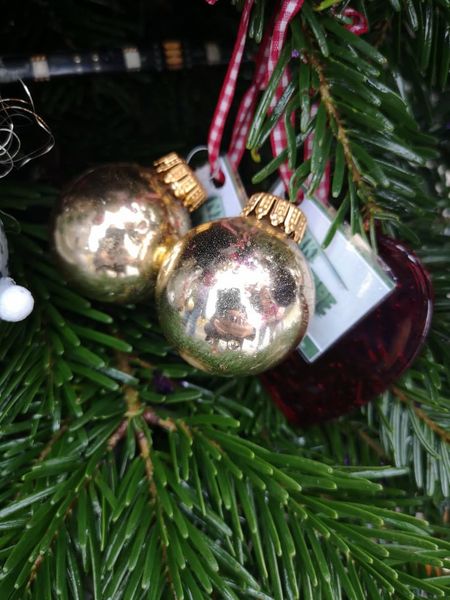 Goldene Weihnachtsbaumkugeln hängen an einem Tannenbaum, umgeben von grünen Nadeln und einem roten Herzanhänger. Die Kugeln reflektieren die Umgebung und verleihen der festlichen Dekoration Glanz.
