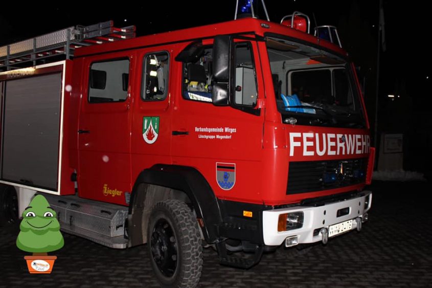 Rotes Feuerwehrfahrzeug der Verbandsgemeinde Wirges mit der Aufschrift "FEUERWEHR", beleuchtet bei Nacht. Das Fahrzeug ist mit einem Leitergestell ausgestattet und zeigt das Wappen der Gemeinde.