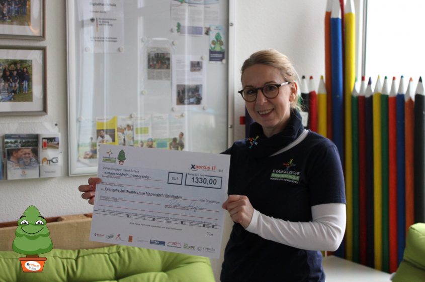 Frau hält einen Scheck über 1330 Euro für die Evangelische Grundschule Mogendorf/Nordhofen, umgeben von bunten Stiften und Informationsmaterialien an der Wand.
