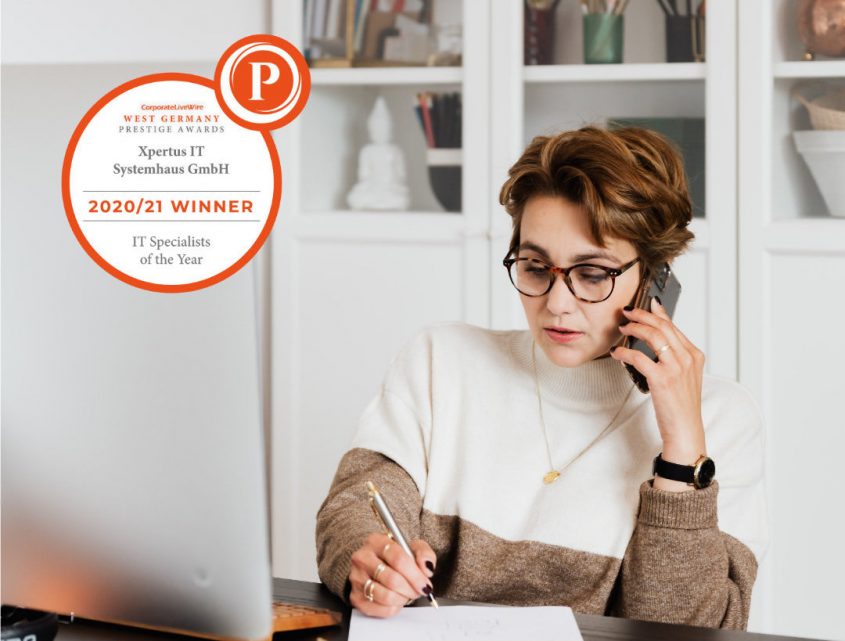 Frau im Büro, die am Telefon spricht und Notizen macht, mit einem Preis für "IT-Spezialisten des Jahres" von Xpertus IT Systemhaus GmbH im Hintergrund.