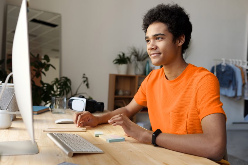 Junger Mann in orangefarbenem Shirt sitzt an einem Schreibtisch und arbeitet am Computer. Auf dem Tisch sind eine Tastatur, ein Maus, einige Holzstäbchen und Notizzettel zu sehen. Im Hintergrund sind Pflanzen und ein Regal mit Kleidung.