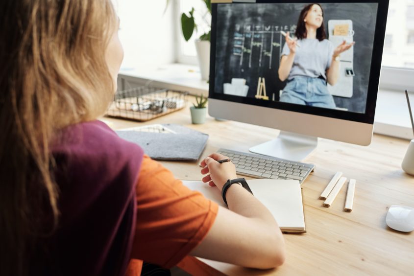 Eine Person sitzt an einem Schreibtisch und schaut auf einen Computerbildschirm, auf dem eine Lehrerin während eines Online-Workshops präsentiert. Auf dem Tisch sind Notizmaterialien und Schreibutensilien sichtbar.