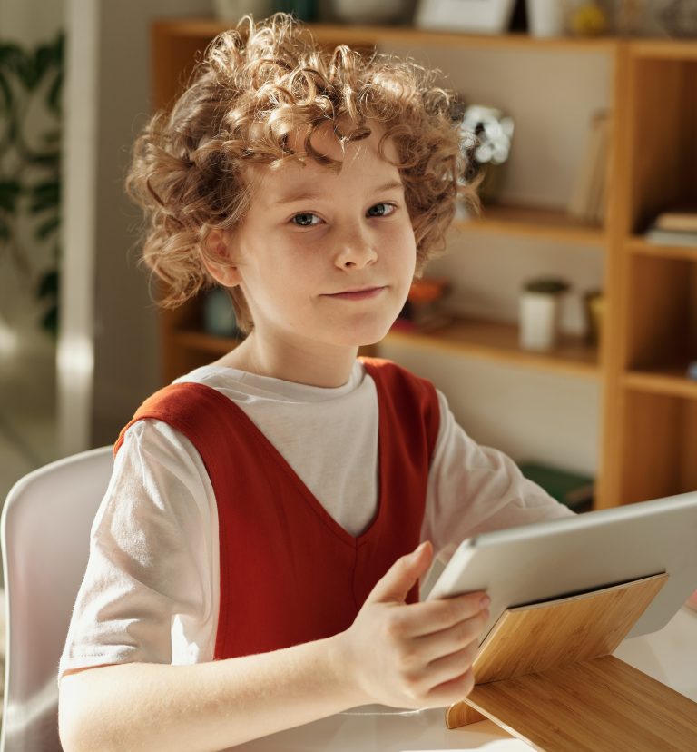 Junge mit lockigem Haar sitzt an einem Tisch und hält ein Tablet in der Hand. Er trägt ein rotes Oberteil über einem weißen T-Shirt. Im Hintergrund sind Regale mit Pflanzen und Dekorationsgegenständen sichtbar.