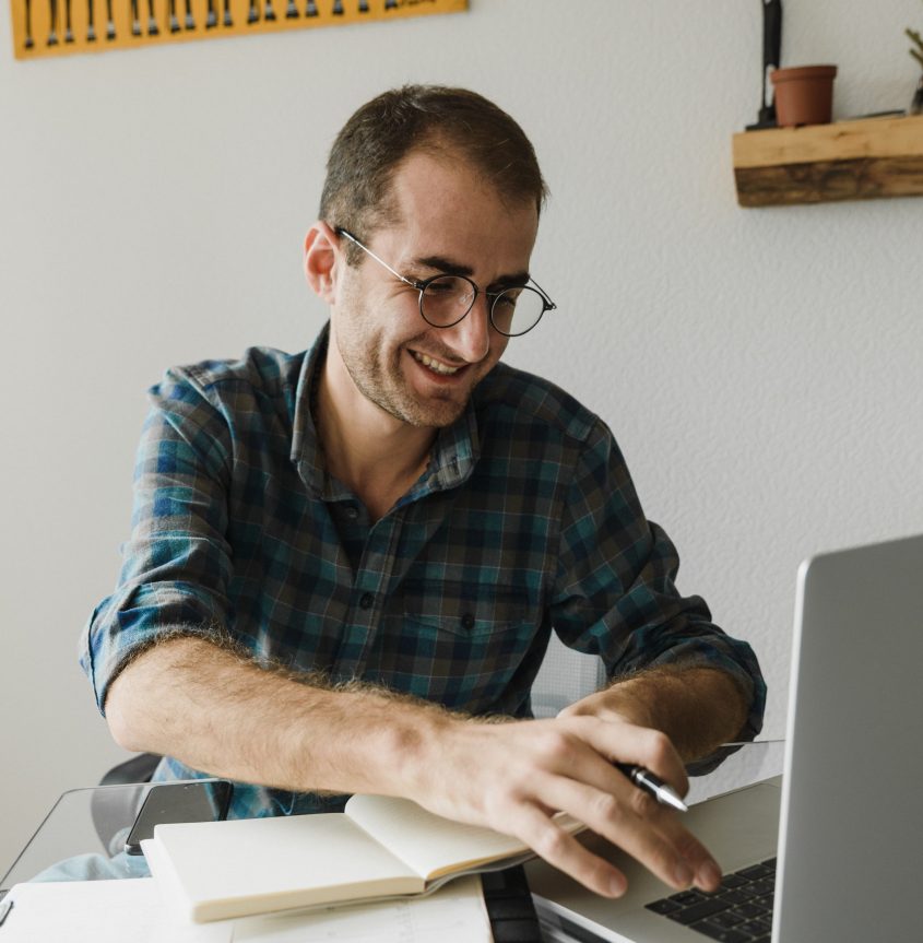 Mann mit Brille und kariertem Hemd, der an einem Laptop arbeitet, lächelt und notiert etwas in einem Notizbuch. Der Arbeitsplatz ist modern und aufgeräumt, mit einer Pflanze im Hintergrund.