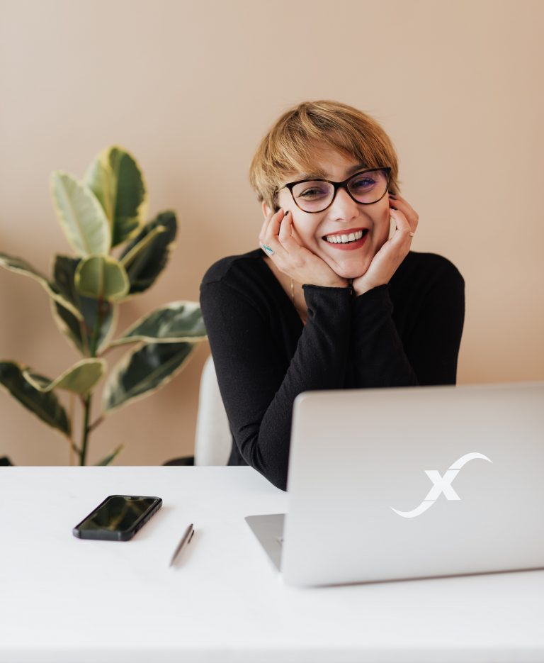 Frau mit Brille lächelt glücklich am Schreibtisch, während sie auf einen Laptop schaut. Neben ihr liegt ein Smartphone und ein Stift. Im Hintergrund ist eine grüne Pflanze vor einer neutralen Wandfarbe zu sehen.