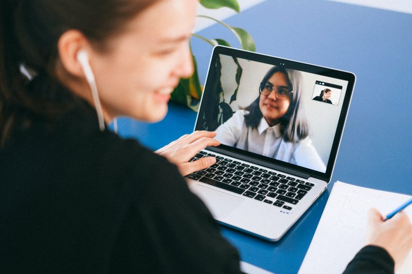 Junge Frau führt ein Videogespräch auf einem Laptop, während sie Notizen macht. Im Hintergrund sind Pflanzen und ein blauer Tisch sichtbar.