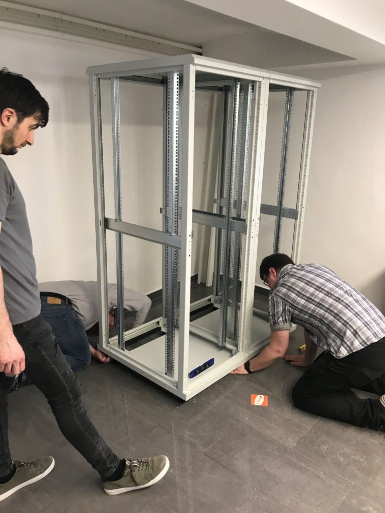 Drei Personen montieren einen Server-Rack in einem Büro. Der Rack ist aus Metall und wird auf einem grauen Boden installiert. Zwei Männer arbeiten am unteren Teil des Racks, während einer den Aufbau überwacht.