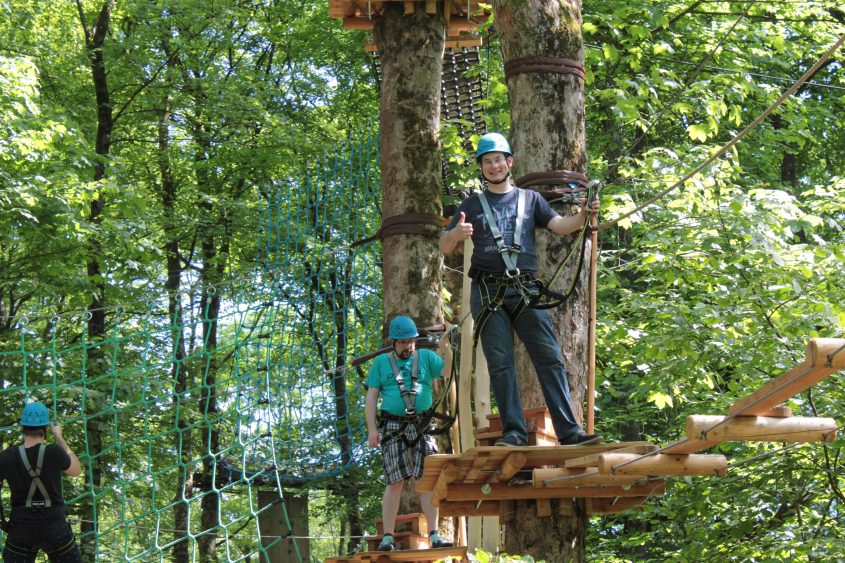 Zwei Männer in Kletterausrüstung stehen auf einer Plattform im Wald, umgeben von Bäumen. Einer der Männer gibt ein Daumen-hoch-Zeichen, während der andere auf einer Netzkonstruktion steht. Die Szene zeigt eine Aktivität im Hochseilgarten.