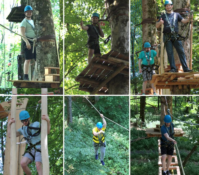 Gruppen von Menschen in einem Kletterpark, die verschiedene Aktivitäten in den Bäumen ausüben, darunter Seilrutschen, Balancieren auf Plattformen und Klettern. Alle tragen Sicherheitshelme und Geschirre. Die Umgebung ist grün und naturnah, ideal für Abenteuer und Outdoor-Aktivitäten.