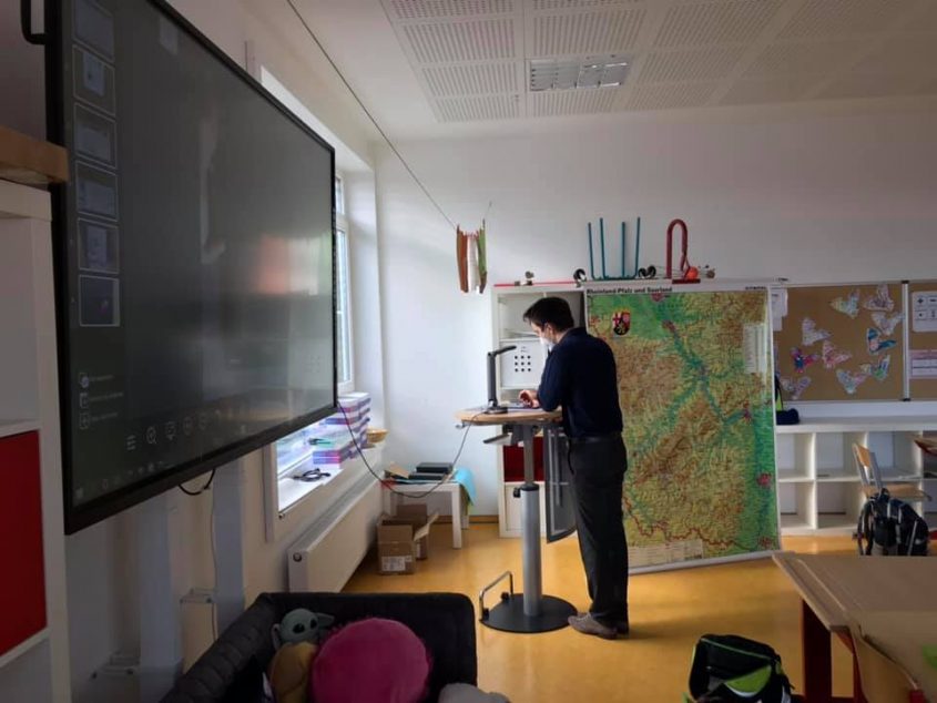 Lehrer steht an einem Stehpult in einem modernen Klassenzimmer, während er mit einem interaktiven Bildschirm und einer großen Karte im Hintergrund arbeitet. Im Raum sind Schulmaterialien und ein gemütlicher Bereich mit Spielzeug sichtbar.