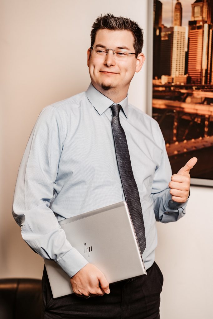 Mann in formeller Kleidung mit Laptop, der lächelt und einen Daumen hoch zeigt, vor einer Bürohintergrundszene.