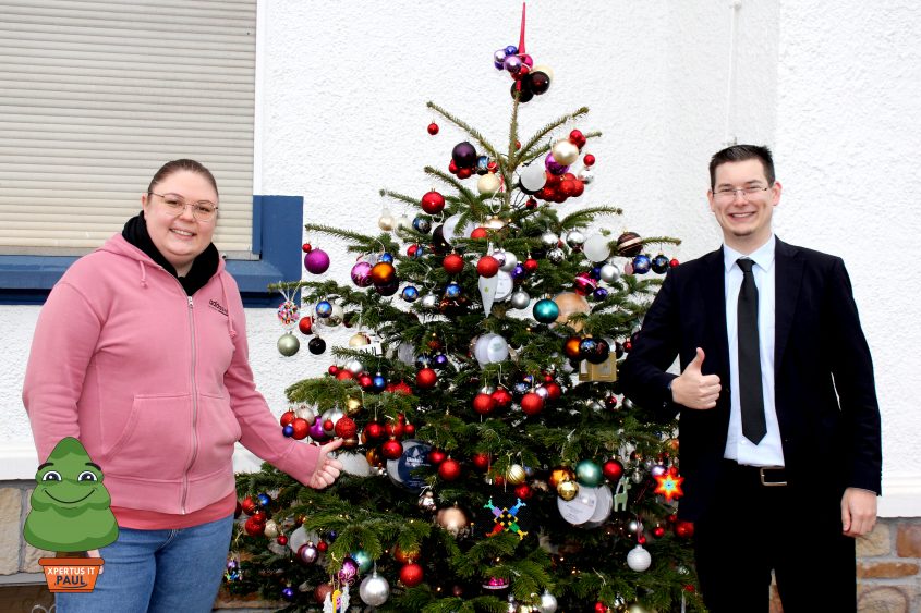 Die zweite große Spendenkugel 35 Frau in rosa Hoodie und Mann im Anzug stehen neben einem festlich geschmückten Weihnachtsbaum mit bunten Kugeln. Der Baum ist reich verziert und die beiden Personen zeigen Daumen hoch, während sie lächeln.