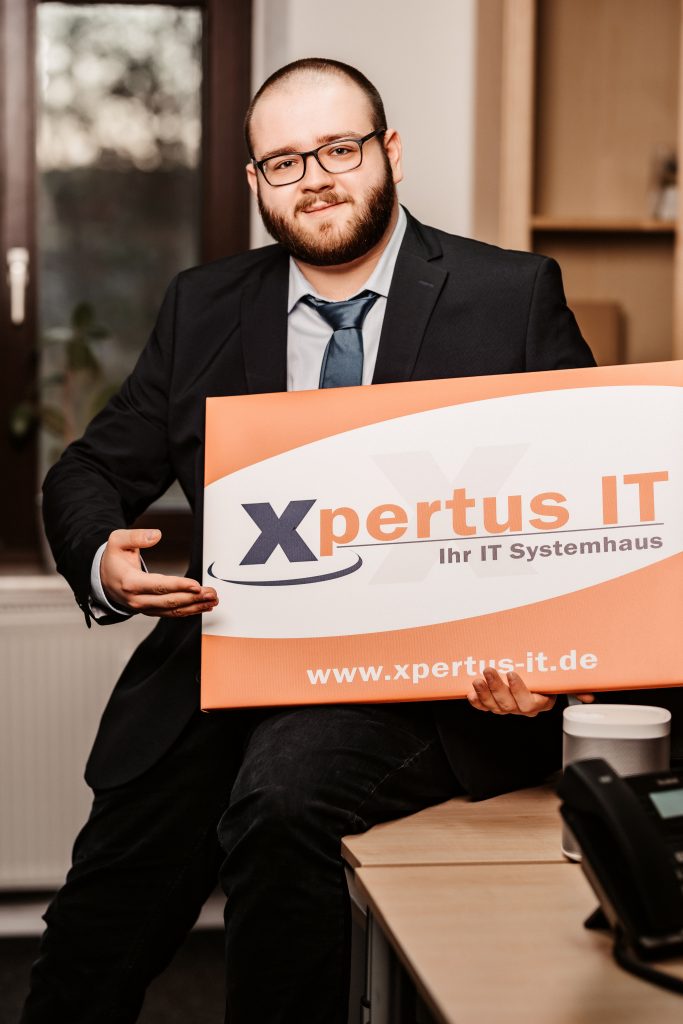 Mann in Anzug sitzt an einem Schreibtisch und hält ein Schild mit dem Logo von Xpertus IT, einem IT-Systemhaus, das Dienstleistungen im Bereich Informationstechnologie anbietet.