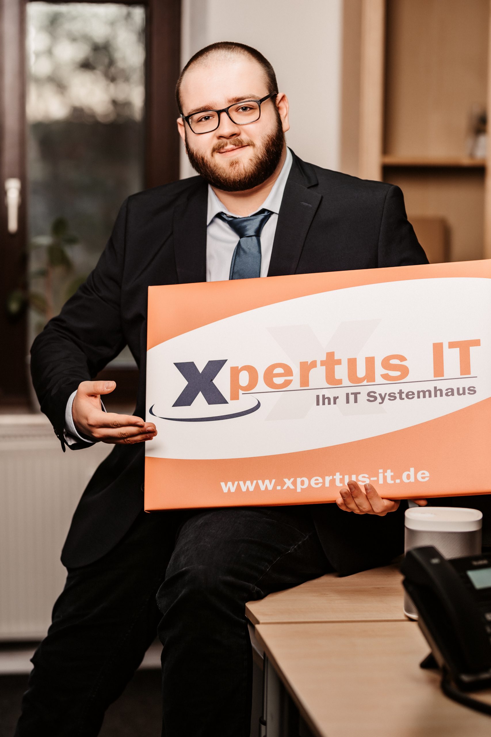 Mann in Anzug sitzt an einem Schreibtisch und hält ein Schild mit dem Logo von Xpertus IT, einem IT-Systemhaus, das Dienstleistungen im Bereich Informationstechnologie anbietet.