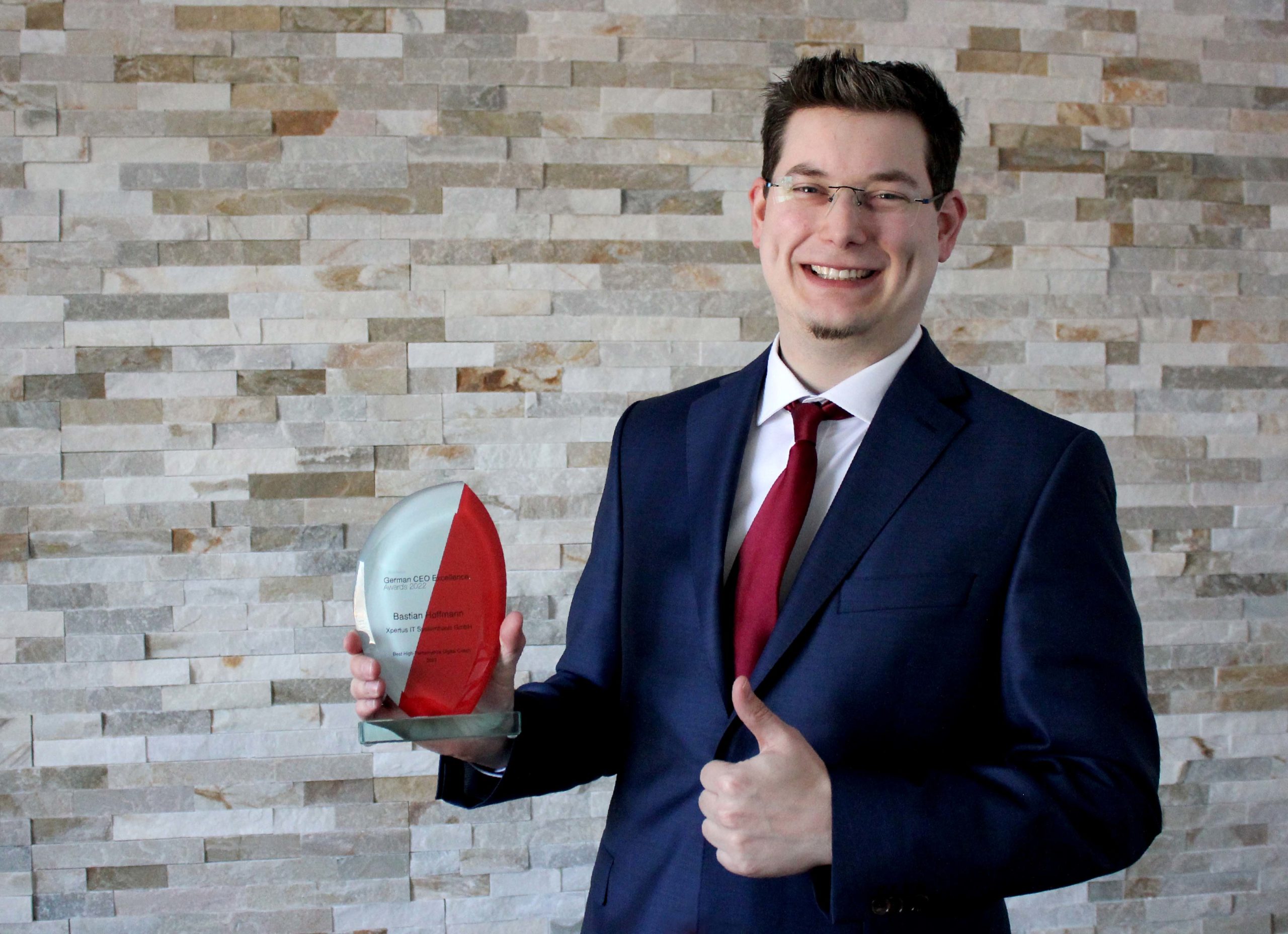 History of Xpertus 15 Mann in Anzug mit roter Krawatte hält einen Preis für herausragende Leistungen in der Unternehmensführung, vor einer modernen Steinwand.