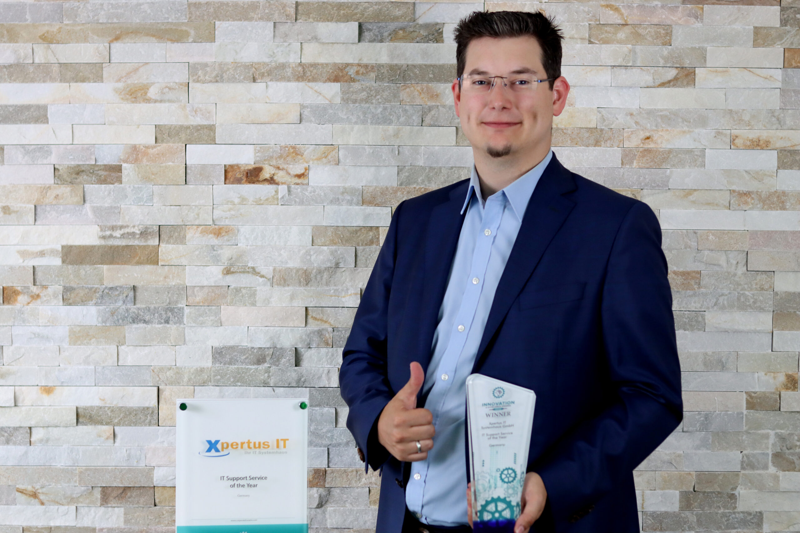 History of Xpertus 12 Mann in Anzug mit Daumen hoch, hält einen Innovationspreis in der Hand, vor einer Steinwand und einem Schild, das die Auszeichnung für den IT-Supportdienst des Jahres von Xpertus IT zeigt.