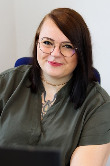 Aktuelles 68 Junge Frau mit Brille und mittellangen, dunklen Haaren, die in einem Büro lächelt. Sie trägt ein grünes Hemd und hat ein auffälliges Tattoo am Hals.