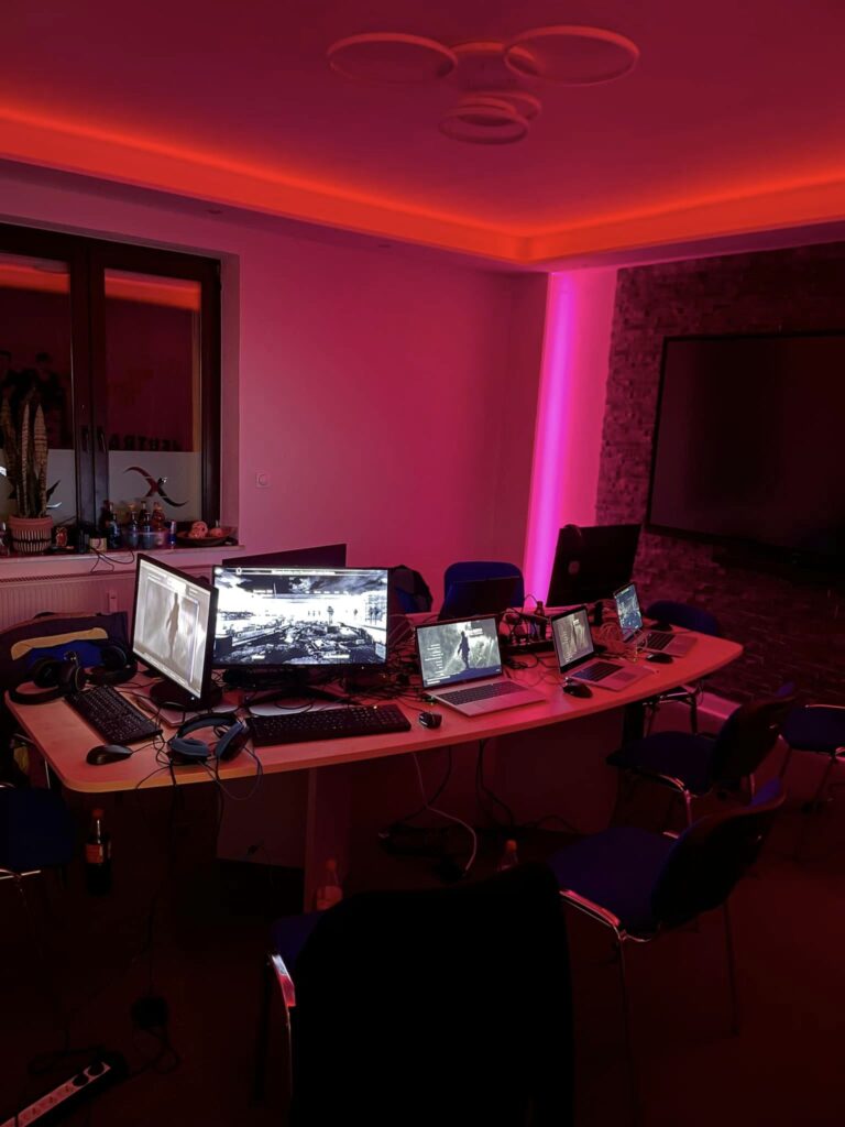 Moderne Gaming-Umgebung mit mehreren Bildschirmen, Laptops und Headsets, beleuchtet von rotem LED-Licht. Der runde Tisch ist umgeben von blauen Stühlen und bietet Platz für eine Gruppe von Gamern.