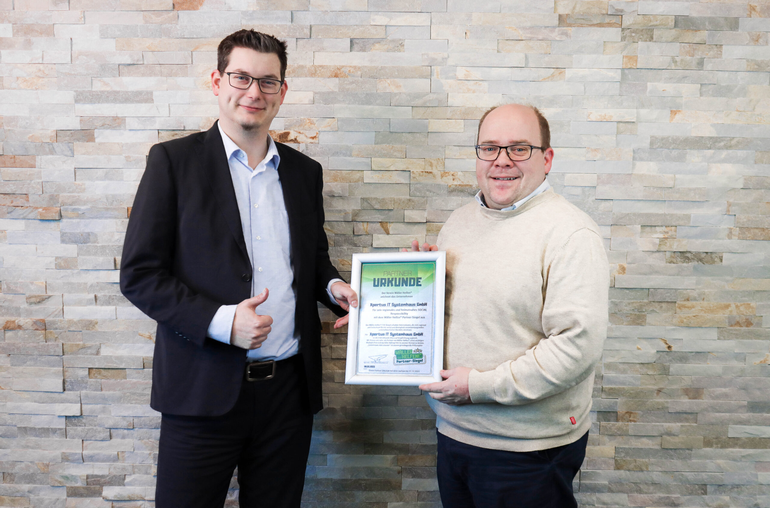 History of Xpertus 9 Zwei Männer stehen vor einer Steinfassade und halten eine Urkunde für die Partnerschaft mit der Firma XY. Der Mann links zeigt einen Daumen nach oben, während der andere lächelt.