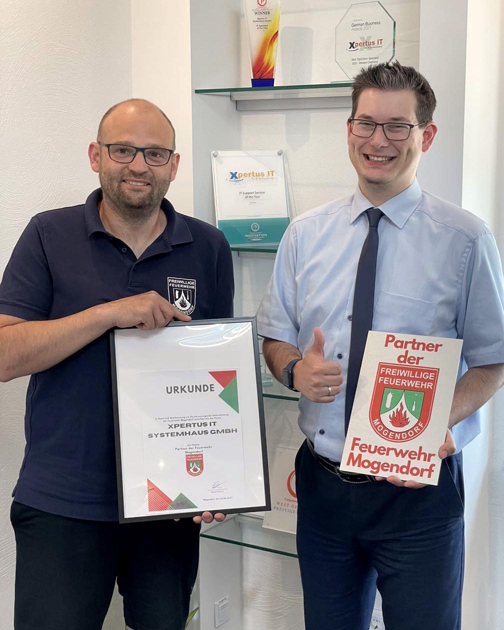 History of Xpertus 6 Zwei Männer stehen in einem Büro und halten Urkunden in den Händen. Der Mann links trägt ein schwarzes T-Shirt mit dem Logo der Freiwilligen Feuerwehr, während der Mann rechts einen blauen Hemd und eine Krawatte trägt. Im Hintergrund sind Auszeichnungen und Zertifikate sichtbar. Die Urkunden bestätigen die Partnerschaft von Xpertus IT Systemhaus GmbH mit der Freiwilligen Feuerwehr Mogendorf.